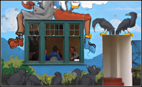 men talking in a coffee shop, seen through a window, with a mural on the wall.